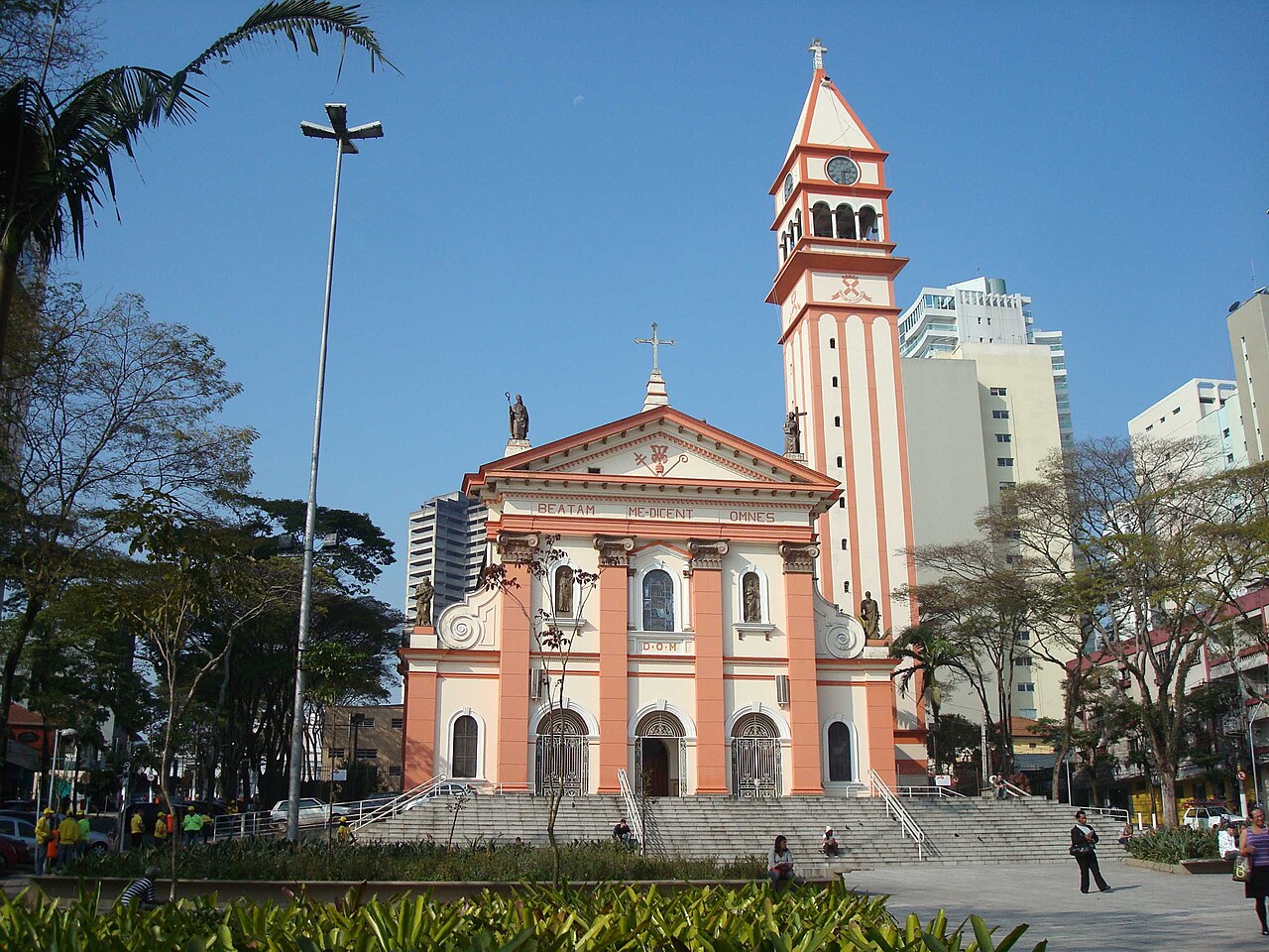 Igreja Matriz de São Bernardo do Campo, com fachada rosa e branca e torre do relógio, em praça arborizada sob céu azul